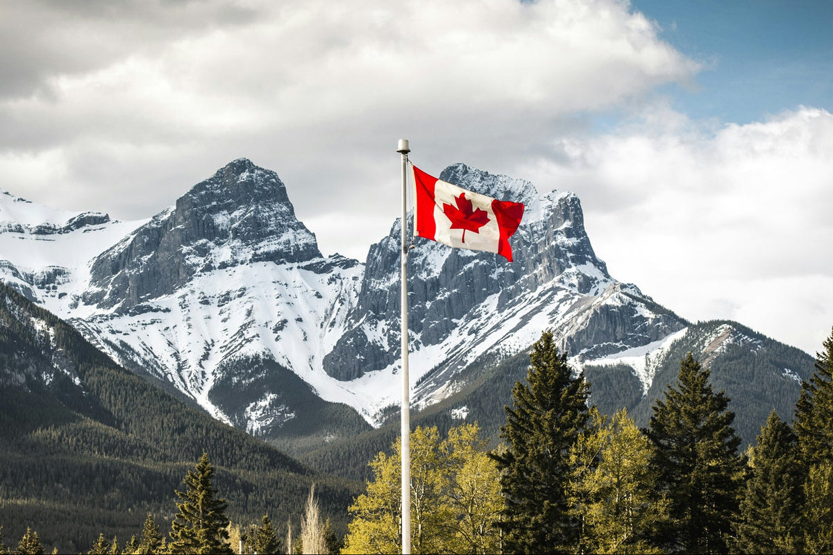 A Canadian flag on a pole waving in front of a dramatic mountain landscape with snow-covered peaks, pine trees, and a partly cloudy blue sky.